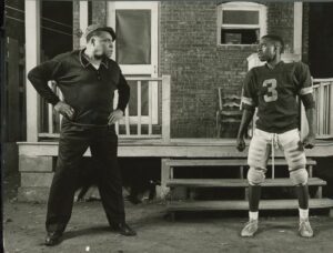 Still from Fences at Yale Repertory Theatre. Two men, including a baseball player in uniform, stand in a confrontational manner in front of a house. 